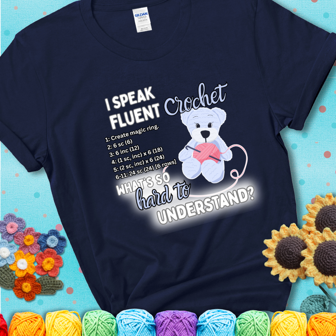 A cute crochet bear holding a yarn ball, surrounded by a short crochet pattern. The phrase reads “I Speak Fluent Crochet – What’s so hard to understand?” on a navy blue t-shirt.