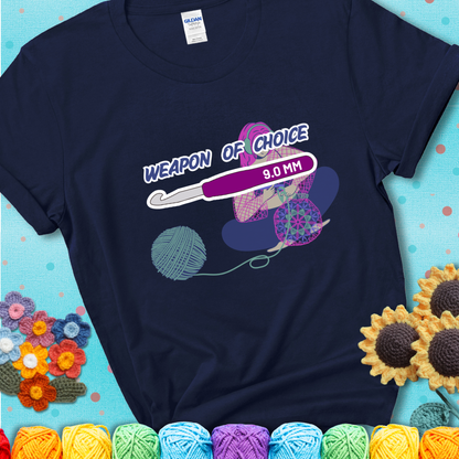 Navy blue t-shirt with an Illustration of a crocheter sitting cross-legged with a 9.0 mm crochet hook above her, labeled “Weapon of Choice.” She’s crocheting a colorful mandala-style project from a large yarn ball.