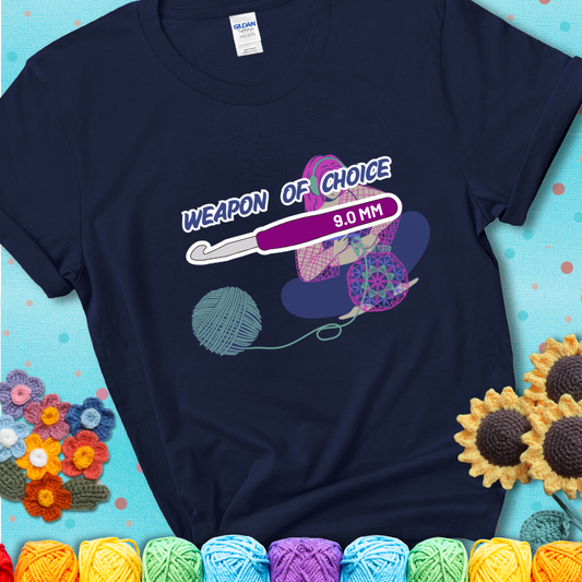 Navy blue t-shirt with an Illustration of a crocheter sitting cross-legged with a 9.0 mm crochet hook above her, labeled “Weapon of Choice.” She’s crocheting a colorful mandala-style project from a large yarn ball.