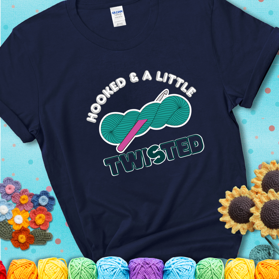A teal skein of yarn with a hot pink crochet hook through it, captioned “Hooked & a Little Twisted” on a navy blue t-shirt.