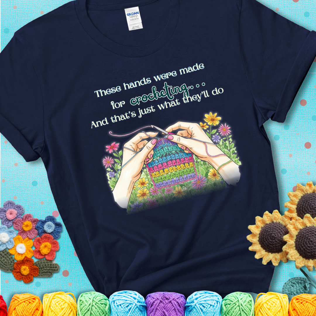 Navy t-shirt with an illustration of two hands crocheting a rainbow swatch, surrounded by colorful flowers and the phrase “These hands were made for crocheting… and that’s just what they’ll do.”