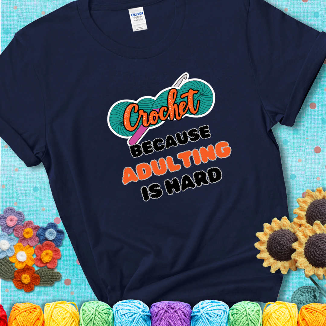 Navy blue t-shirt featuring a bold design with the phrase “Crochet Because Adulting Is Hard” featuring a green skein of yarn with a pink crochet hook. The word “Crochet” is in a script font, while “Adulting is hard” is in chunky textured letters.