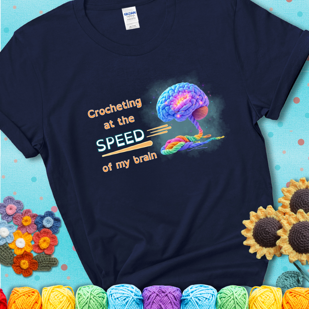 A glowing rainbow brain unravels into colorful yarn with the phrase “Crocheting at the speed of my brain” on a navy blue t-shirt. 