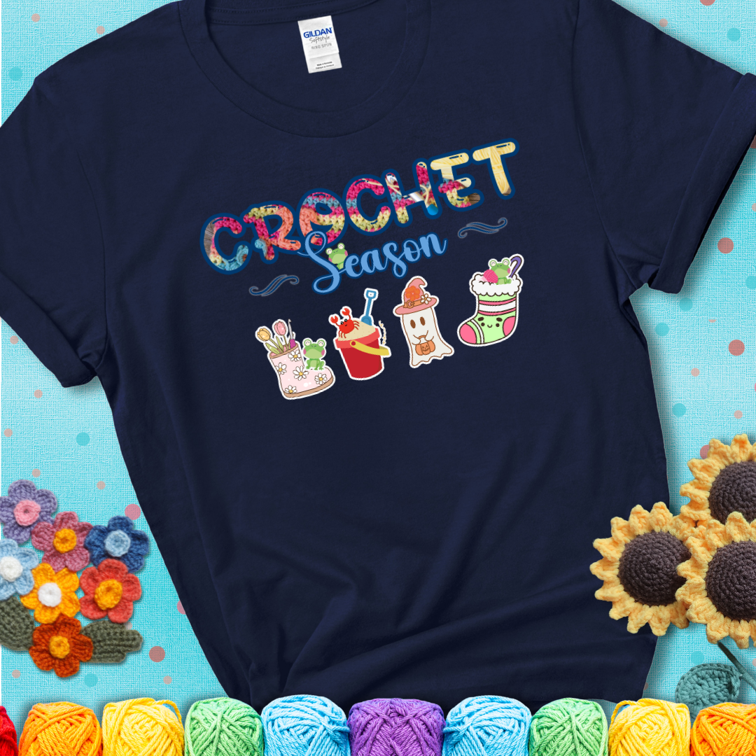 Navy blue t-shirt shows the words “Crochet Season” with cute seasonal icons below: a spring boot with hooks, summer bucket with crab, fall ghost with pumpkin hat, winter stocking, and tiny frogs.