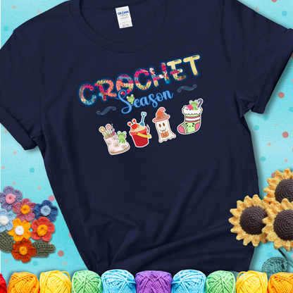 Navy blue t-shirt shows the words “Crochet Season” with cute seasonal icons below: a spring boot with hooks, summer bucket with crab, fall ghost with pumpkin hat, winter stocking, and tiny frogs.
