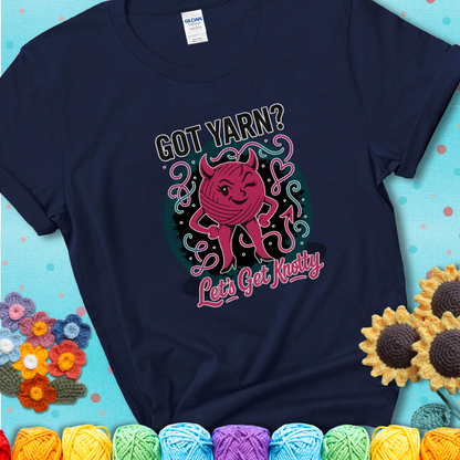 Navy blue t-shirt with a cheeky pink yarn ball wearing devil horns, arms, and legs, winking beneath the phrase “Got Yarn? Let’s Get Knotty