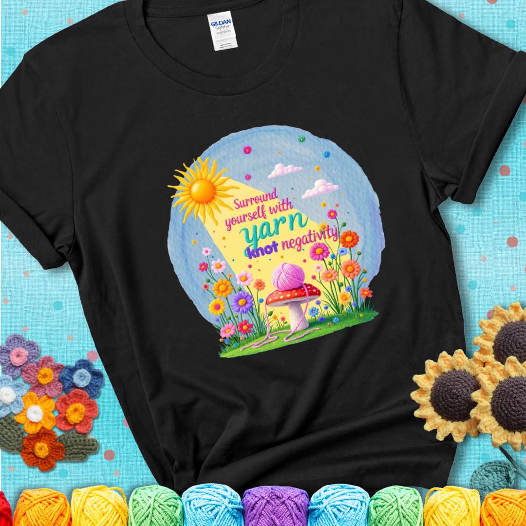 Black  t-shirt featuring a happy spring garden scene with a sunbeam, flowers, and a pink yarn ball on a mushroom, with the quote "Surround yourself with yarn, knot negativity."