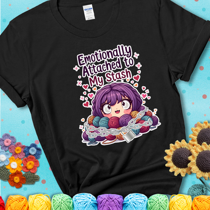 Adorable character with wide eyes hugging a yarn stash wrapped in chains and a lock, with the text “Emotionally Attached to My Stash”  on a black tee.