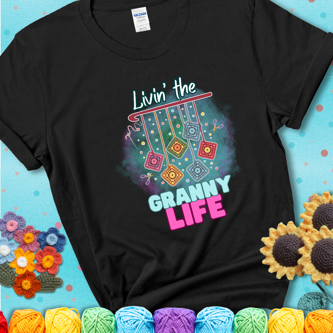 Black t-shirt shows colorful crochet granny squares hanging from a hook with text “Livin’ the Granny Life” in bold pink and blue font; a fun nod to classic crochet.
