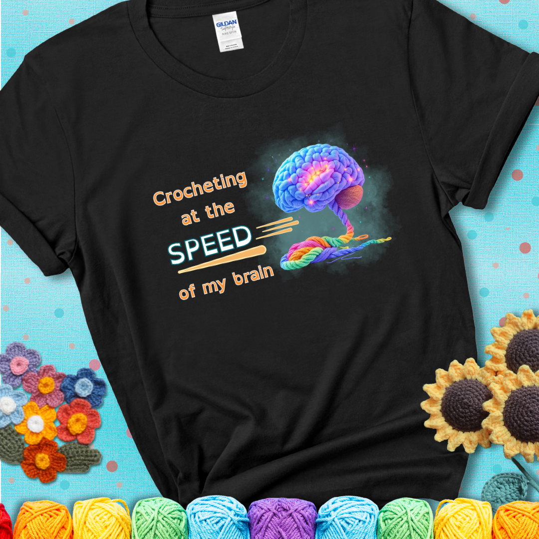 A glowing rainbow brain unravels into colorful yarn with the phrase “Crocheting at the speed of my brain” on a black t-shirt. 