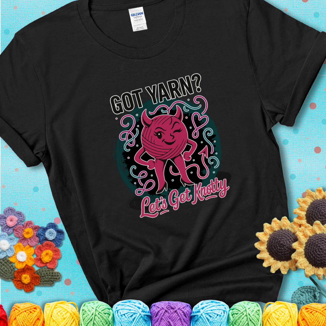 Black t-shirt with a cheeky pink yarn ball wearing devil horns, arms, and legs, winking beneath the phrase “Got Yarn? Let’s Get Knotty