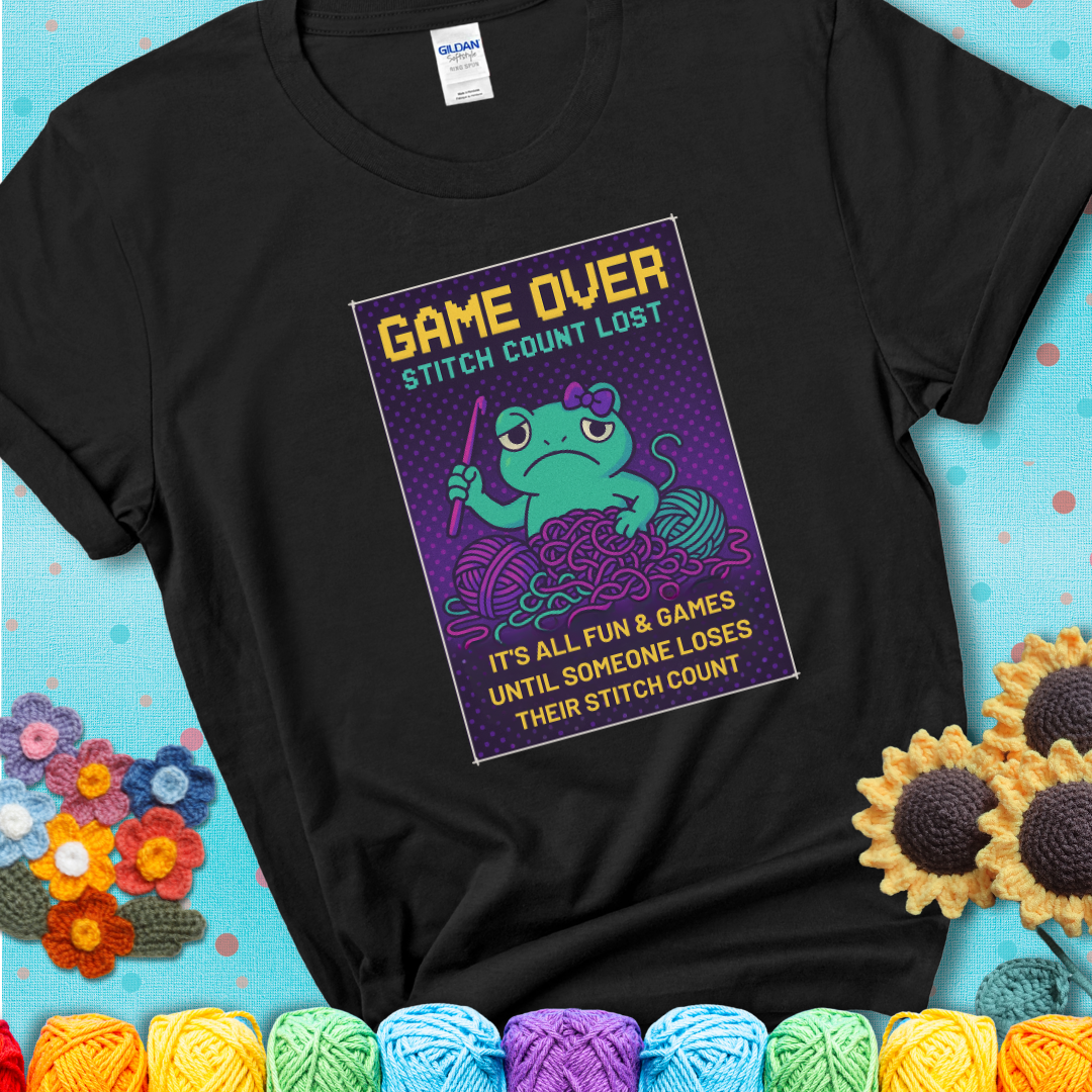 Black t-shirt shows a pixel-style design of a grumpy yarn frog holding a hook, surrounded by yarn chaos, with the text “Game Over. Stitch Count Lost.”