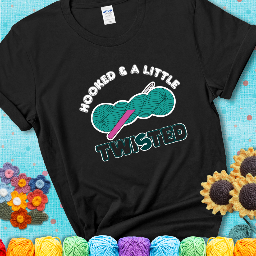 A teal skein of yarn with a hot pink crochet hook through it, captioned “Hooked & a Little Twisted” on a black t-shirt.