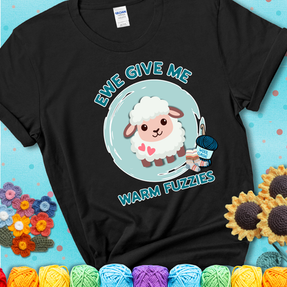 Black t-shirt showing a cartoon sheep with pink hearts, wool socks, and yarn labeled “Wool Classic.” Text reads “Ewe Give Me Warm Fuzzies.”