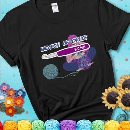 Black t-shirt with an Illustration of a crocheter sitting cross-legged with a 9.0 mm crochet hook above her, labeled “Weapon of Choice.” She’s crocheting a colorful mandala-style project from a large yarn ball.
