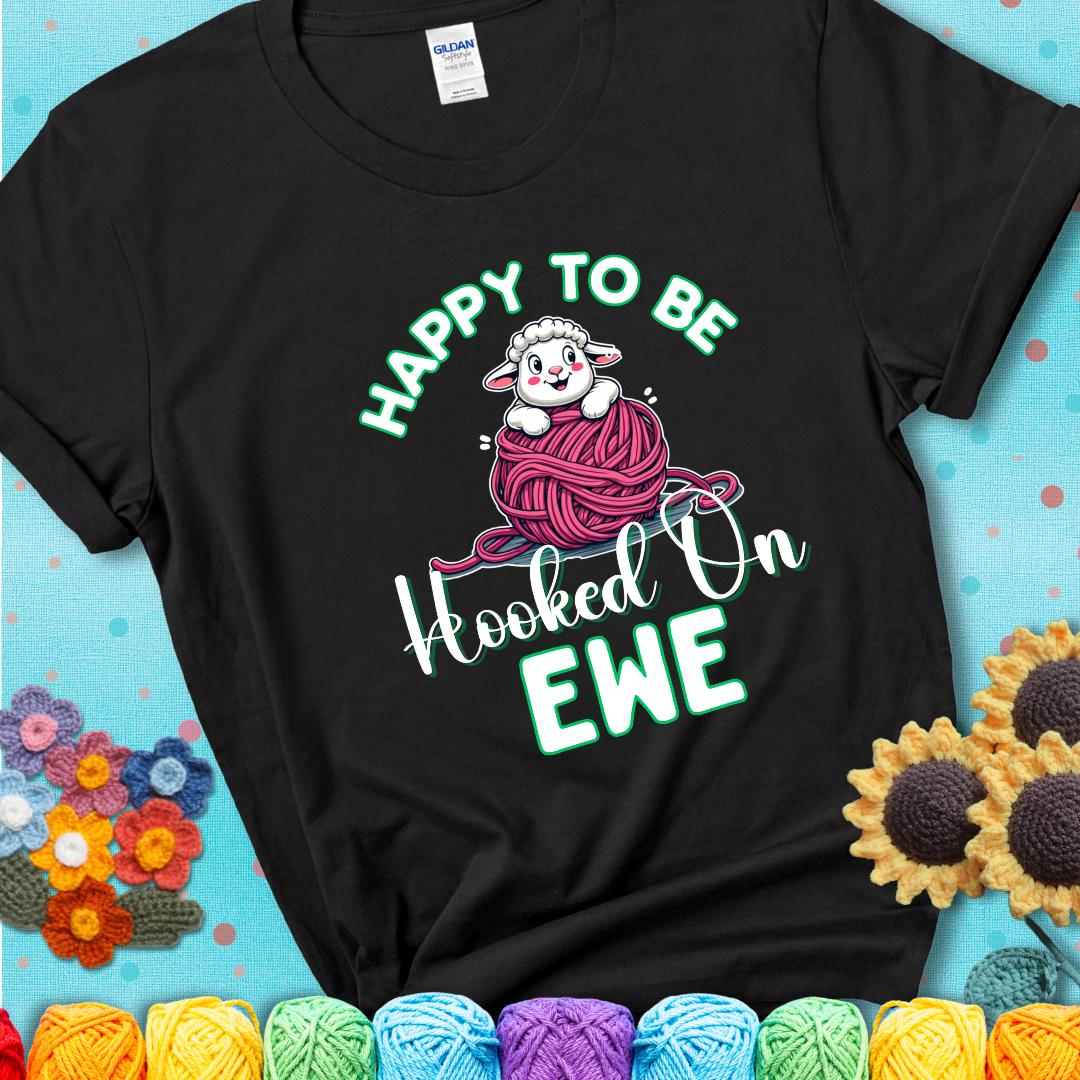 Black t-shirt with smiling cartoon sheep peeks from a pink yarn ball, surrounded by green text “Happy to Be Hooked on Ewe”, a playful and affectionate sheep pun for crocheters.