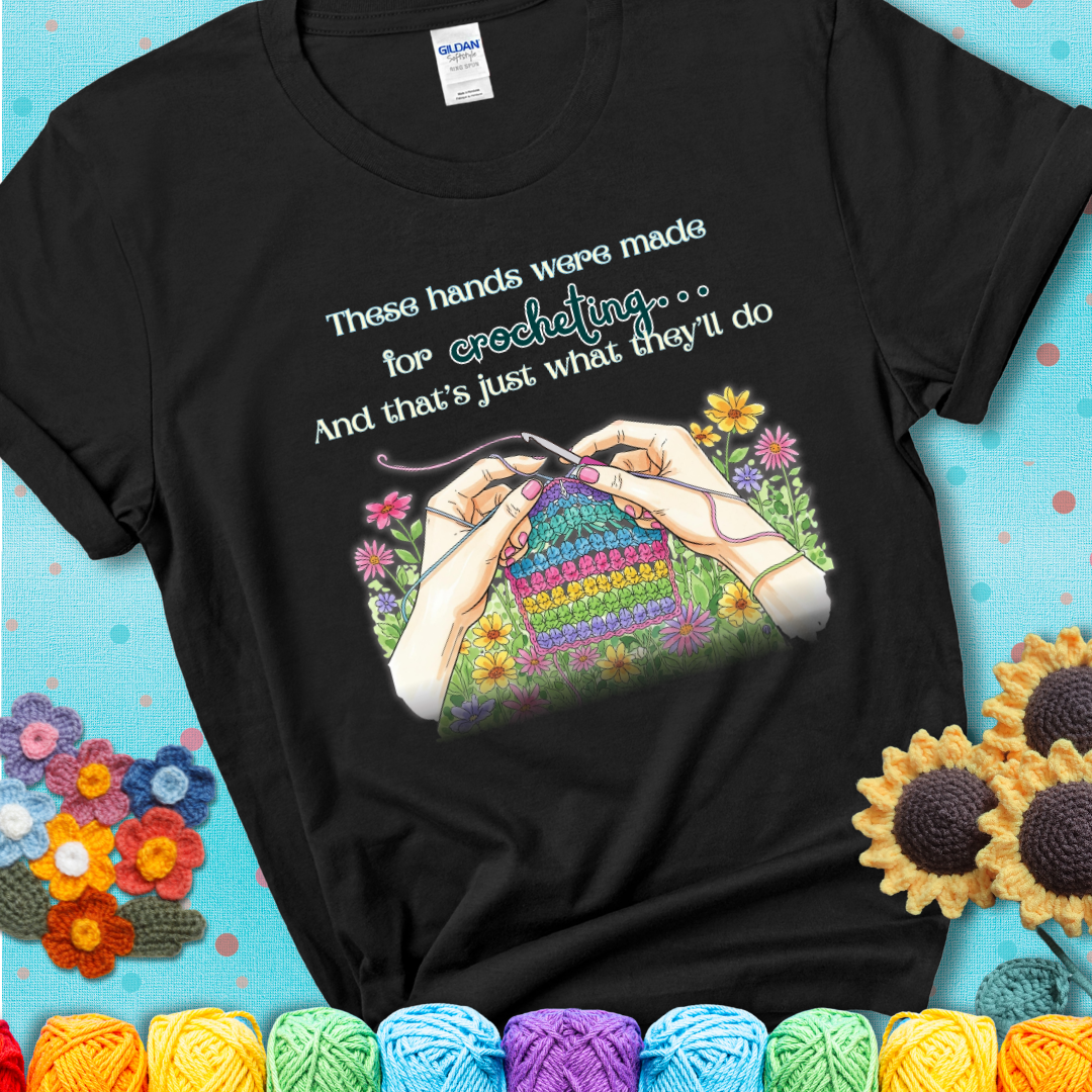 Black t-shirt with an illustration of two hands crocheting a rainbow swatch, surrounded by colorful flowers and the phrase “These hands were made for crocheting… and that’s just what they’ll do.”