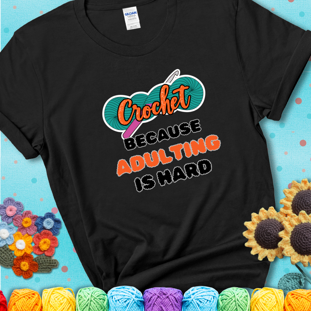 Black t-shirt featuring a bold design with the phrase “Crochet Because Adulting Is Hard” featuring a green skein of yarn with a pink crochet hook. The word “Crochet” is in a script font, while “Adulting is hard” is in chunky textured letters.