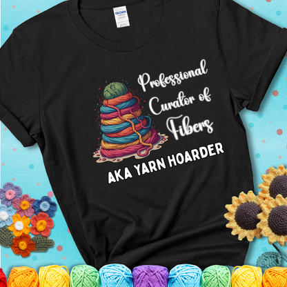 A black t-shirt showing a pyramid of rainbow yarn topped with green ball; text reads “Professional Curator of Fibers AKA Yarn Hoarder”, a playful shirt for serious stashers.