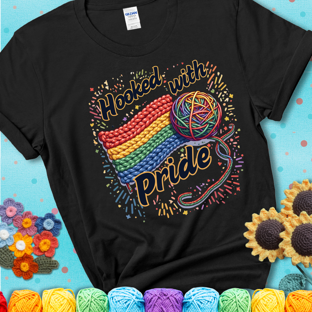 A rainbow-striped crochet swatch paired with a multicolor yarn ball and bold “Hooked With Pride” lettering. Confetti surrounds the vibrant scene showing a black tee with a teal sunflower background
