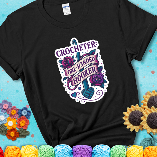Black t-shirt with a tattoo-style crochet design featuring a crochet hook, roses, and the phrase “Crocheter: One-Handed Hooker.”