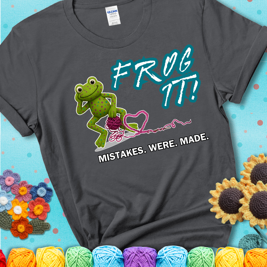 Green frog with yarn and heart-shaped loop. Text reads: “Frog It! Mistakes Were Made.” A playful crochet tee for anyone who's ever had to rip it back, showing a charcoal grey tee with a teal sunflower background
