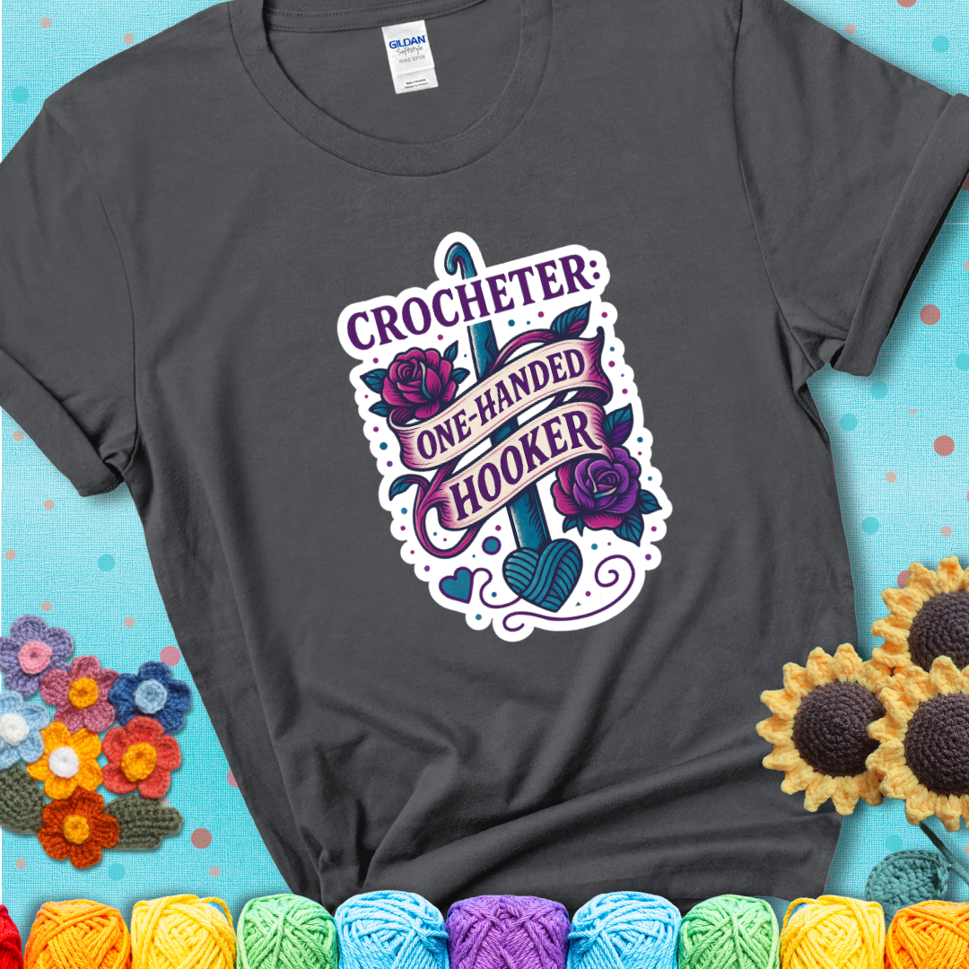Charcoal t-shirt with a tattoo-style crochet design featuring a crochet hook, roses, and the phrase “Crocheter: One-Handed Hooker.”