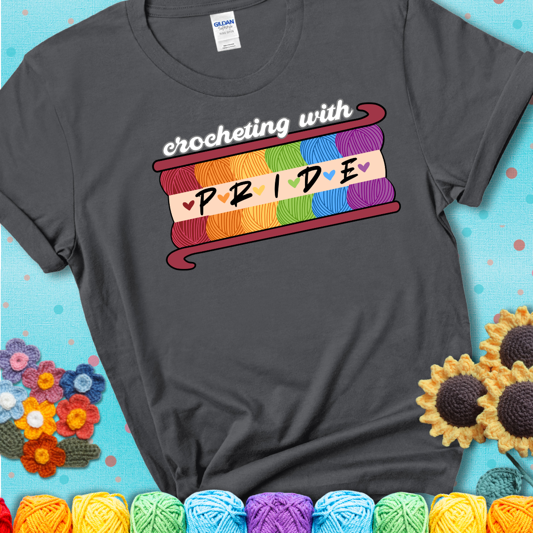 Yarn skeins in rainbow colors stacked in a hook shape. Text reads “Crocheting With Pride” showing a charcoal grey tee with a teal sunflower background