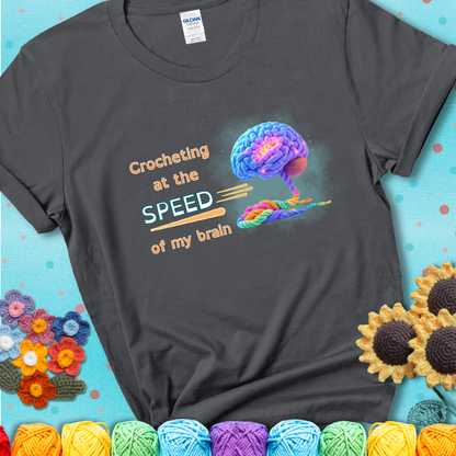 A glowing rainbow brain unravels into colorful yarn with the phrase “Crocheting at the speed of my brain” on a charcoal grey t-shirt. 
