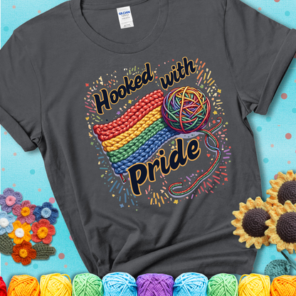 A rainbow-striped crochet swatch paired with a multicolor yarn ball and bold “Hooked With Pride” lettering. Confetti surrounds the vibrant scene showing a charcoal grey tee with a teal sunflower background