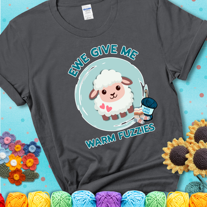 Charcoal grey t-shirt showing a cartoon sheep with pink hearts, wool socks, and yarn labeled “Wool Classic.” Text reads “Ewe Give Me Warm Fuzzies.”