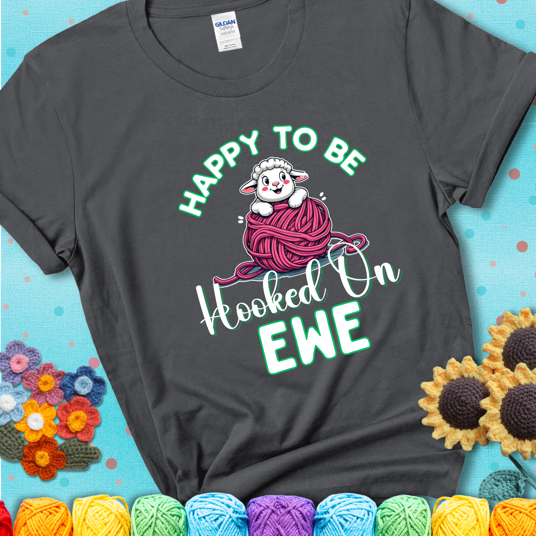 Charcoal grey t-shirt with smiling cartoon sheep peeks from a pink yarn ball, surrounded by green text “Happy to Be Hooked on Ewe”, a playful and affectionate sheep pun for crocheters.