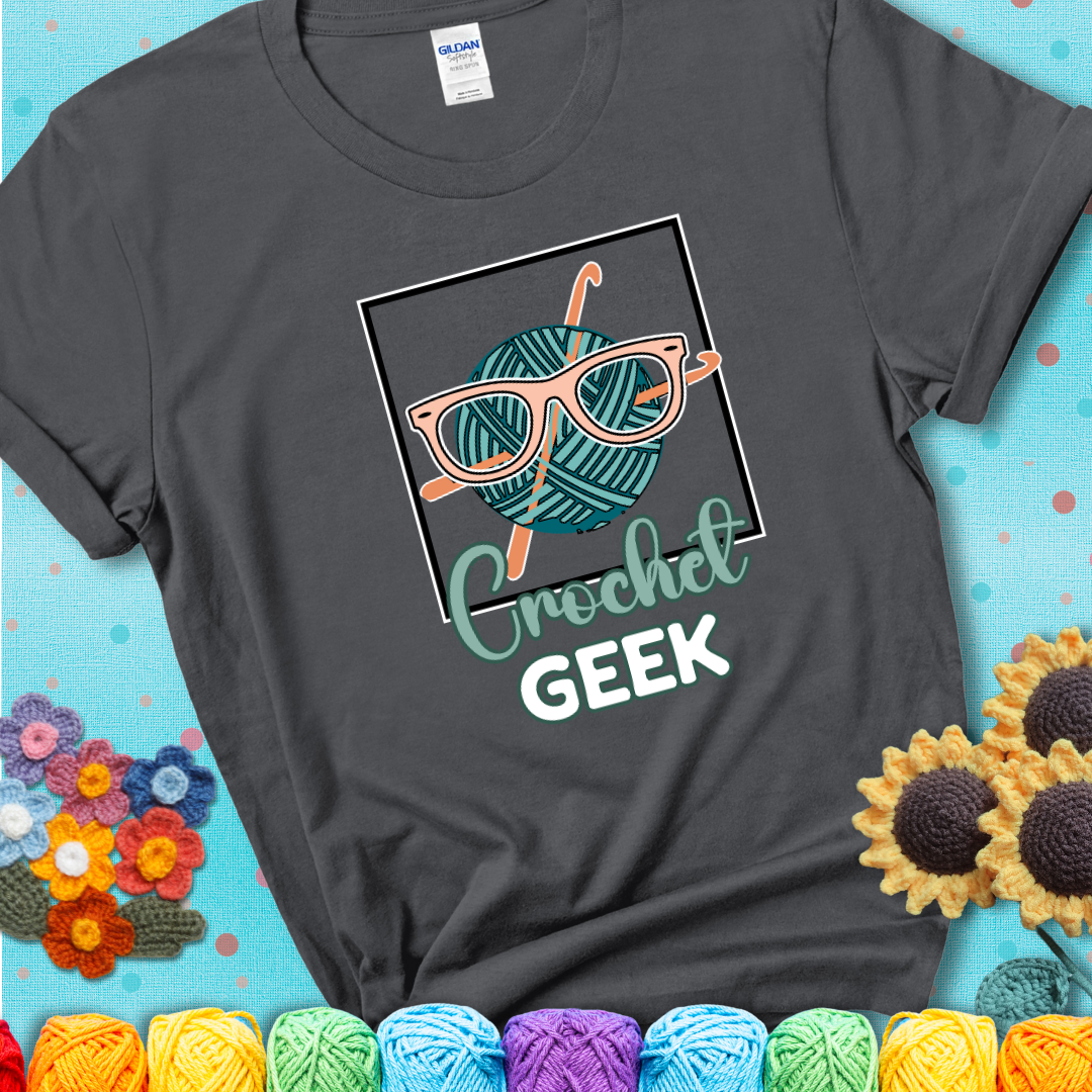 A teal yarn ball with crisscrossed hooks wears oversized nerdy glasses with the phrase “Crochet Geek” in bold, fun text below on a charcoal grey t-shirt.