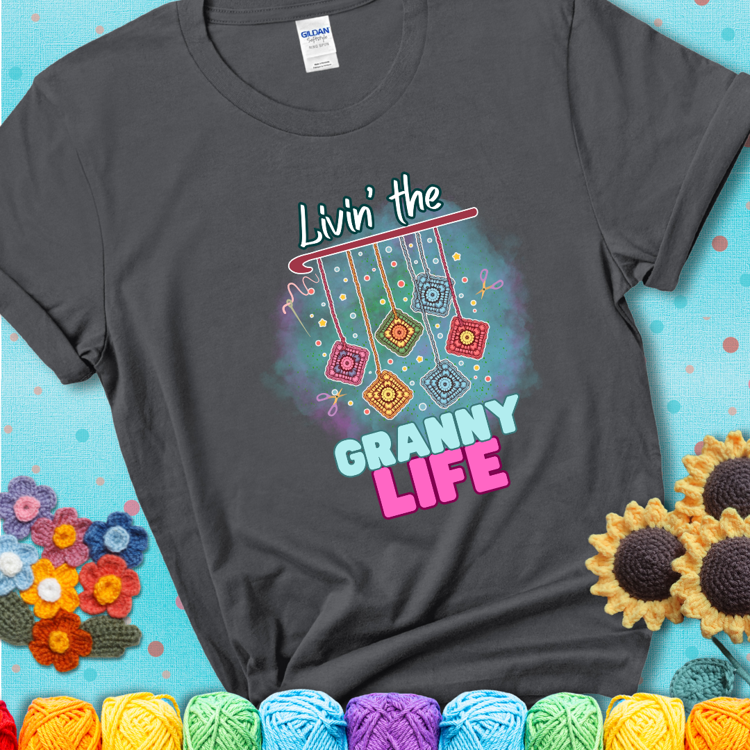 Charcoal grey t-shirt shows colorful crochet granny squares hanging from a hook with text “Livin’ the Granny Life” in bold pink and blue font; a fun nod to classic crochet.
