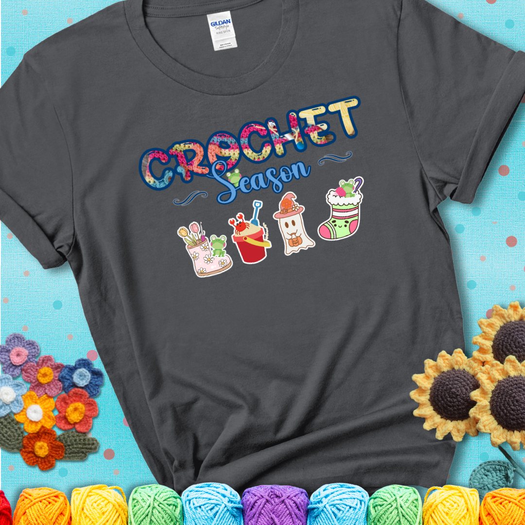 Charcoal grey t-shirt shows the words “Crochet Season” with cute seasonal icons below: a spring boot with hooks, summer bucket with crab, fall ghost with pumpkin hat, winter stocking, and tiny frogs.