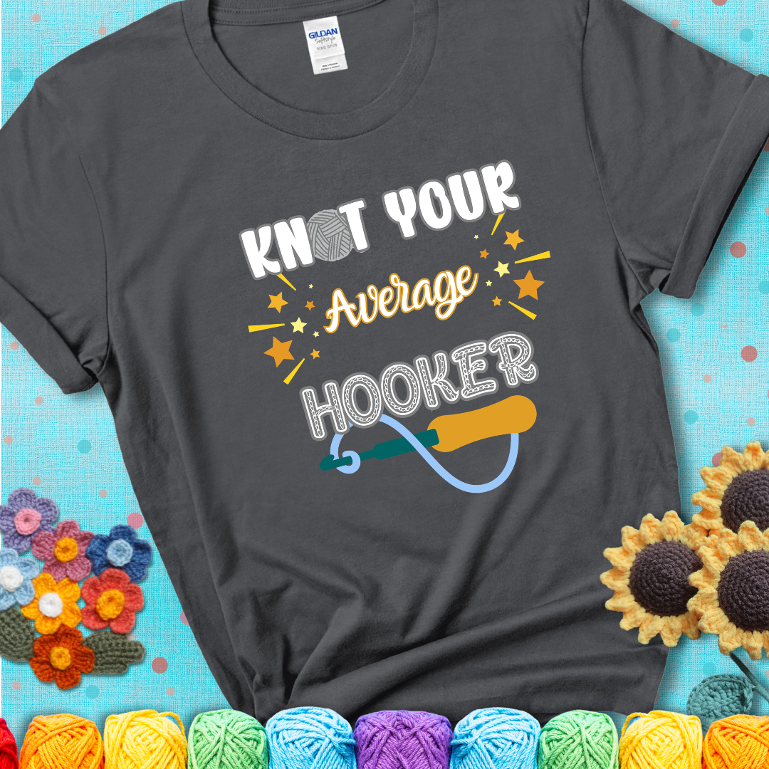 Bold lettering on a charcoal grey t-shirt with a yarn hook graphic. Text reads: “I'm Not Your Average Hooker.” A cheeky crochet tee for confident crafters who own their skills.