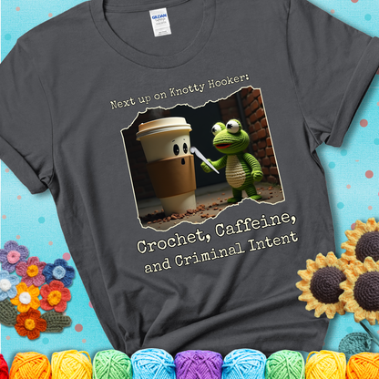A crocheted frog holding a crochet hook confronts a surprised coffee cup in a dim alleyway with text: "Next up on Knotty Hooker: Crochet, Caffeine, and Criminal Intent," showing a charcoal grey tee with a teal sunflower background