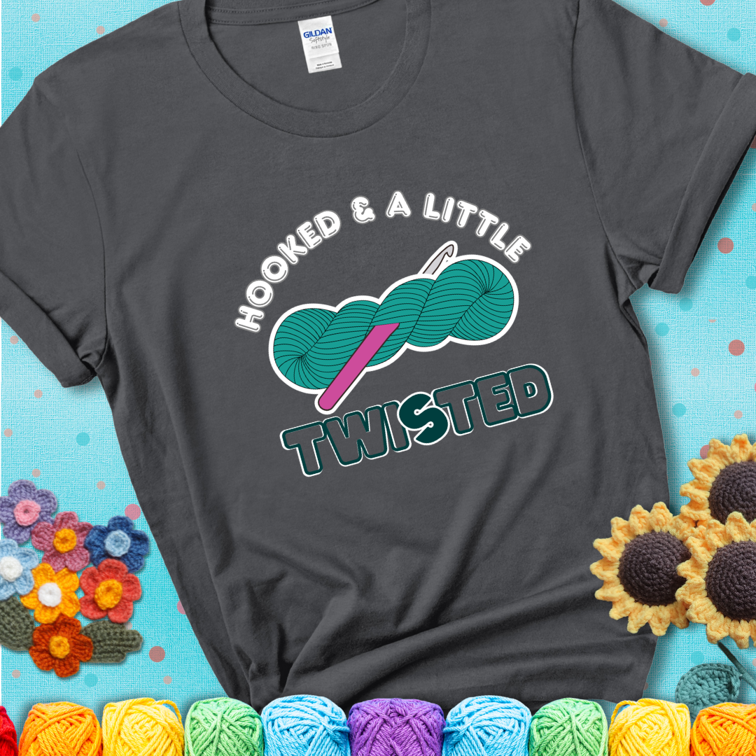 A teal skein of yarn with a hot pink crochet hook through it, captioned “Hooked & a Little Twisted” on a charcoal grey t-shirt.