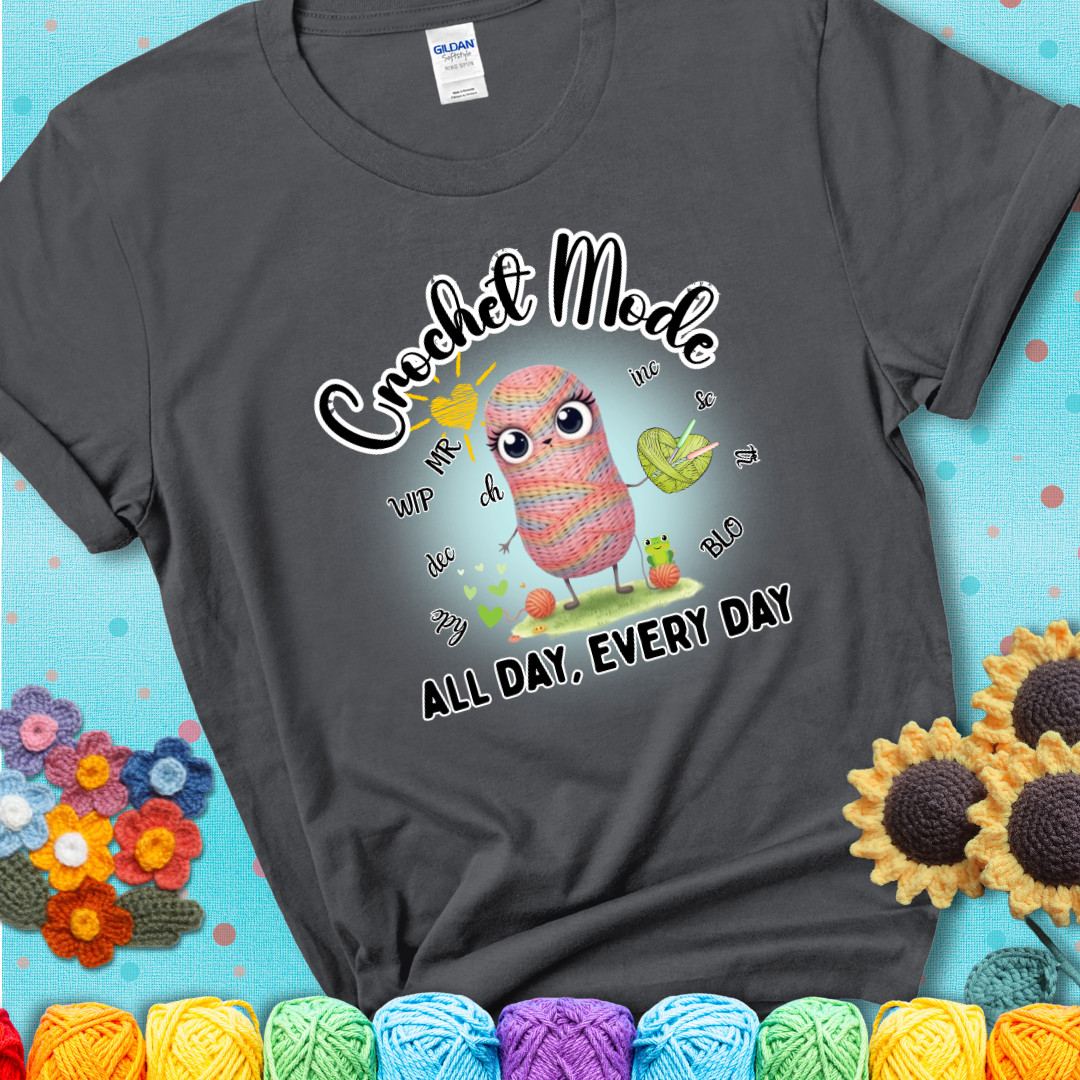 Charcoal grey t-shirt with a kawaii-style rainbow yarn character surrounded by crochet stitch abbreviations and holding a yarn heart, with text “Crochet Mode All Day, Every Day.”