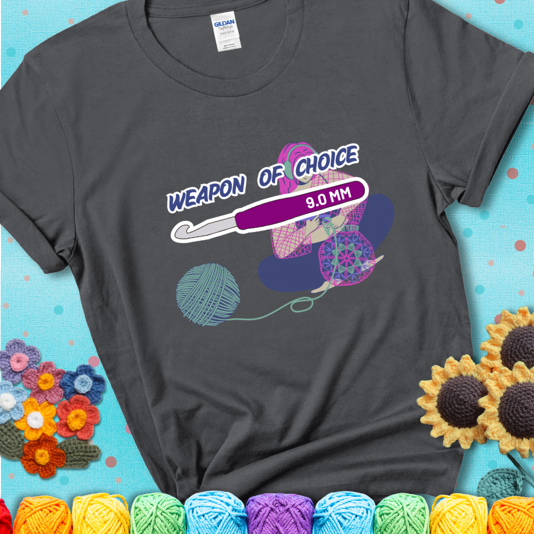 Charcoal grey t-shirt with an Illustration of a crocheter sitting cross-legged with a 9.0 mm crochet hook above her, labeled “Weapon of Choice.” She’s crocheting a colorful mandala-style project from a large yarn ball.