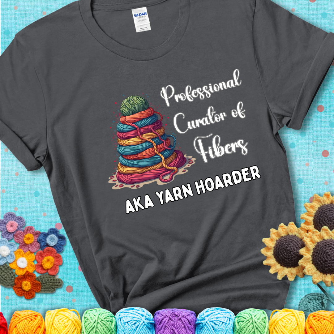 A charcoal grey t-shirt showing a pyramid of rainbow yarn topped with green ball; text reads “Professional Curator of Fibers AKA Yarn Hoarder”, a playful shirt for serious stashers.