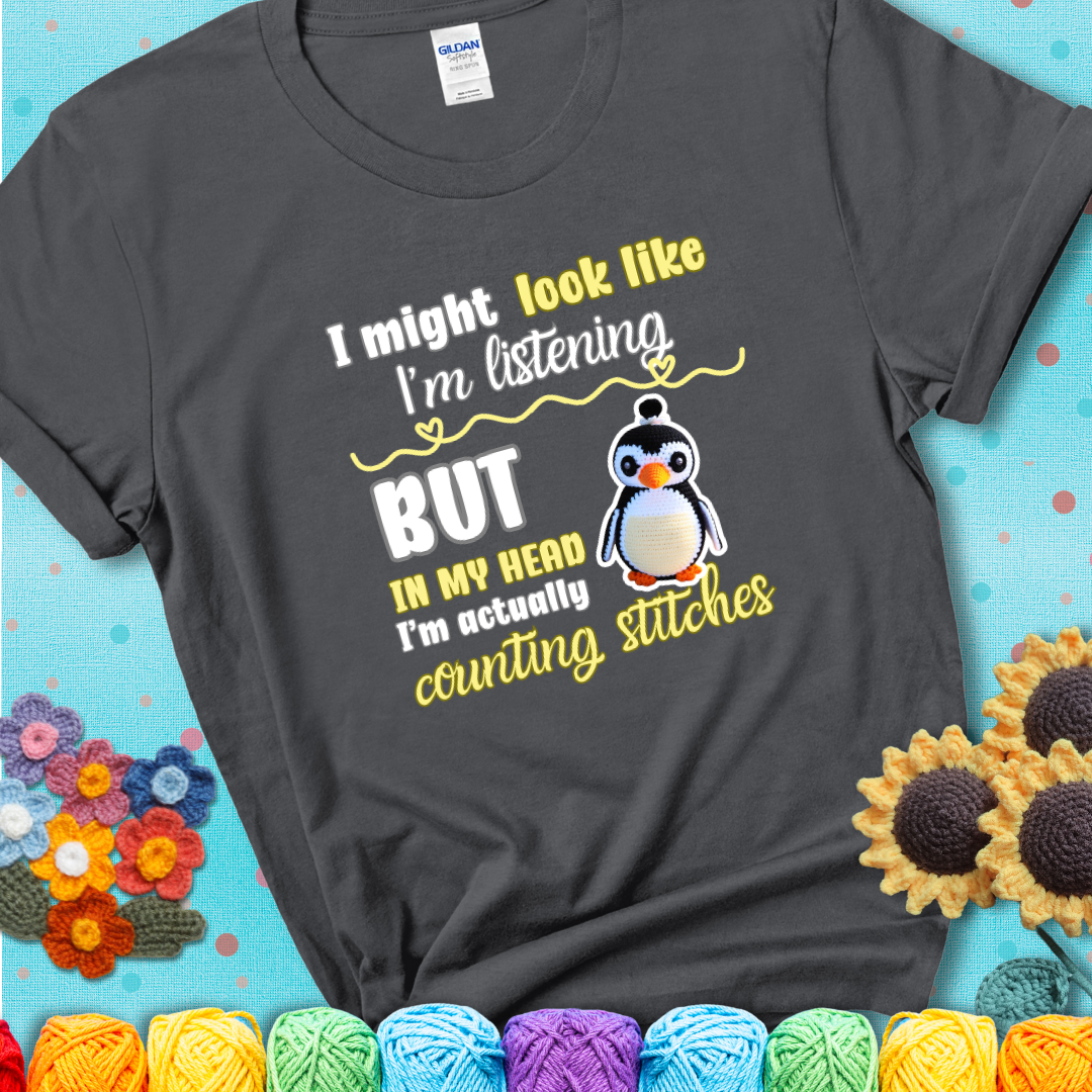 Charcoal grey t-shirt shows a crocheted penguin next to the text “I might look like I’m listening but in my head I’m actually counting stitches.”