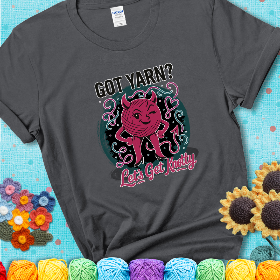 Charcoal t-shirt with a cheeky pink yarn ball wearing devil horns, arms, and legs, winking beneath the phrase “Got Yarn? Let’s Get Knotty