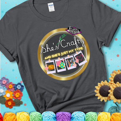 Text on charcoal grey tee reads "She’s Crafty & She’s Just My Type" with yarn, fabric, painting, and cross-stitch icons hanging like Polaroids