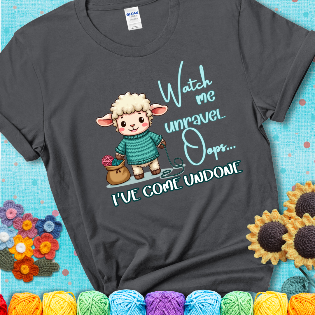 Charcoal grey t-shirt with a cute sheep holding yarn looks startled with the phrase “Watch me unravel… Oops, I’ve come undone.” Perfect mix of whimsy and yarn life chaos.