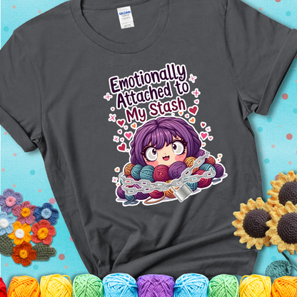 Adorable character with wide eyes hugging a yarn stash wrapped in chains and a lock, with the text “Emotionally Attached to My Stash”  on a charcoal grey tee.