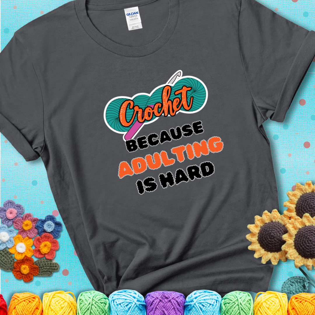 Charcoal grey t-shirt featuring a bold design with the phrase “Crochet Because Adulting Is Hard” featuring a green skein of yarn with a pink crochet hook. The word “Crochet” is in a script font, while “Adulting is hard” is in chunky textured letters.
