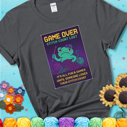 Charcoal grey t-shirt shows a pixel-style design of a grumpy yarn frog holding a hook, surrounded by yarn chaos, with the text “Game Over. Stitch Count Lost.”
