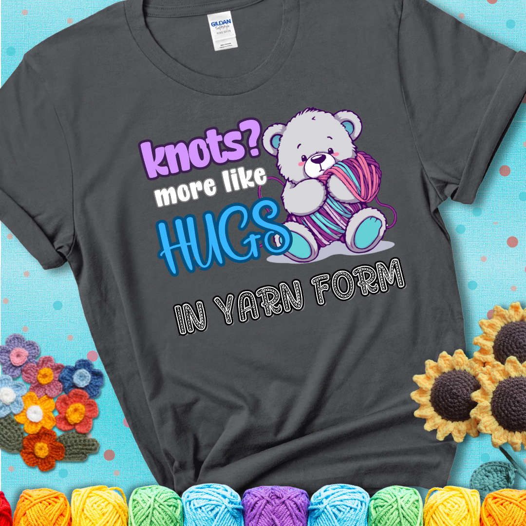 Cute teddy bear holding colorful yarn ball with hearts. Text reads: “Knots? More Like Hugs in Yarn Form.” Perfect for cozy gift-givers and yarn lovers, showing a charcoal grey tee with a teal sunflower background