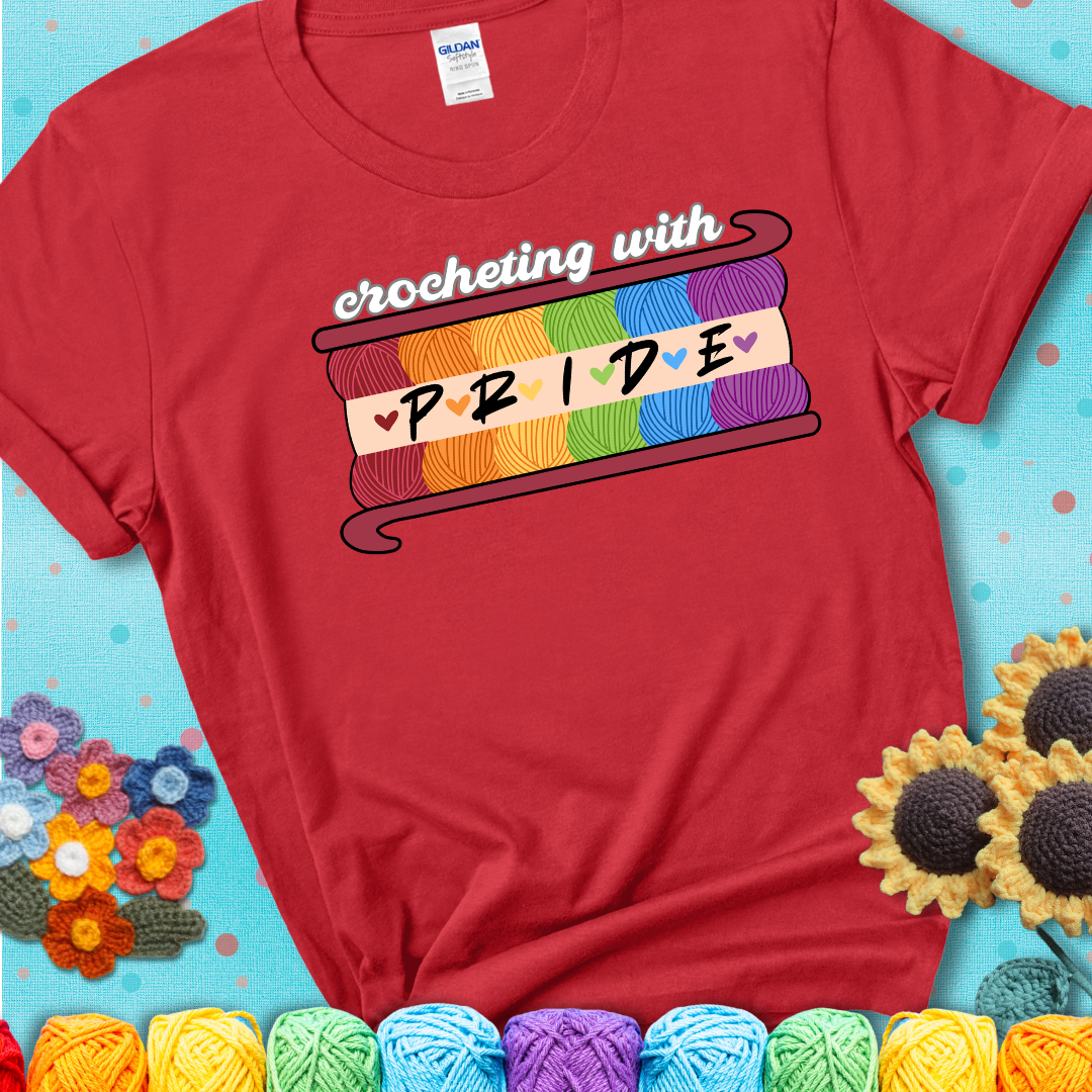 Yarn skeins in rainbow colors stacked in a hook shape. Text reads “Crocheting With Pride” showing a red tee with a teal sunflower background